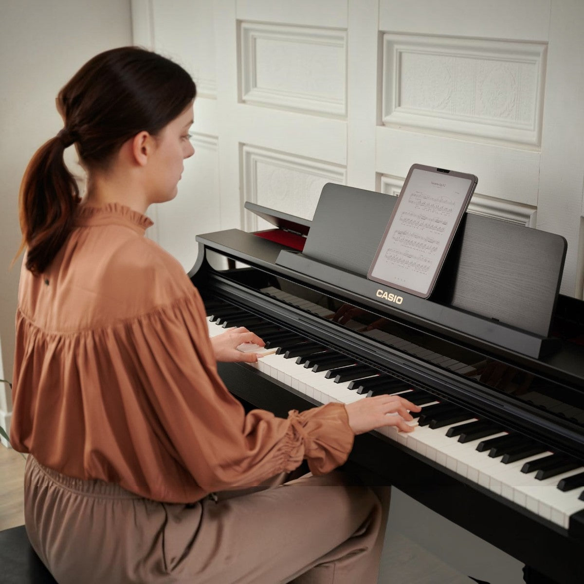 Người phụ nữ đang chơi đàn piano điện Casio AP-550 màu đen tại Tân Nhạc Cụ với máy tính bảng đặt trên giá nhạc