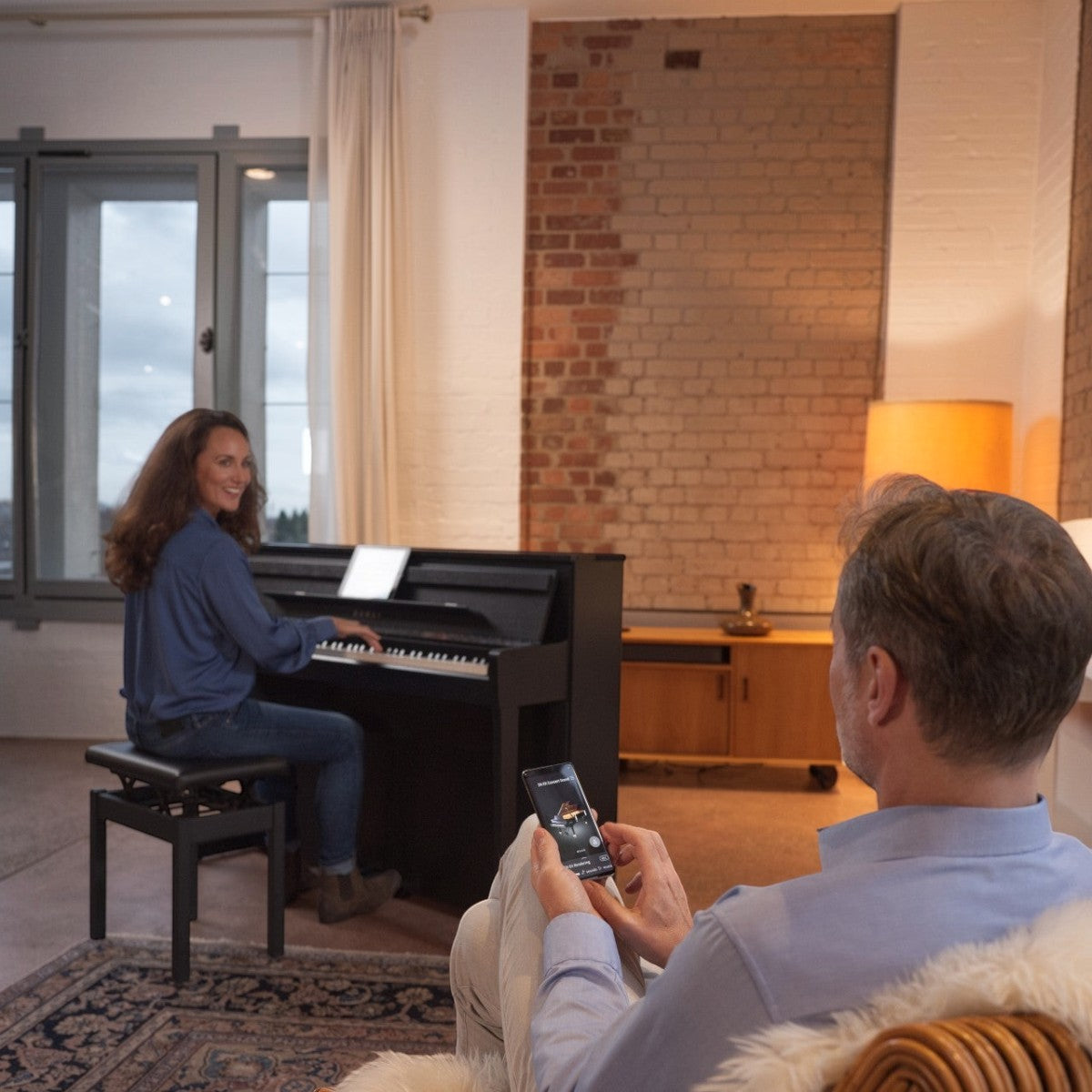 Người phụ nữ chơi đàn piano điện Kawai CA99 tại Tân Nhạc Cụ trong khi người đàn ông điều khiển qua ứng dụng điện thoại