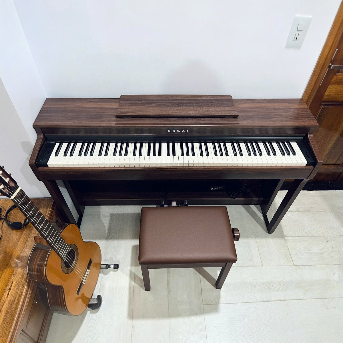 Đàn piano điện Kawai CN201 dáng tủ màu gỗ tại Tân Nhạc Cụ, kèm ghế ngồi và đàn guitar bên cạnh