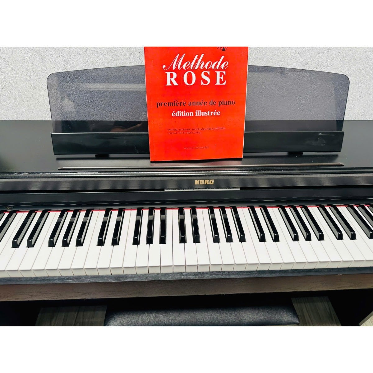 Đàn piano điện TÂN NHẠC CỤ Korg C2000 màu gỗ nâu đen với nắp trượt và sách nhạc Methode Rose đặt trên giá đỡ