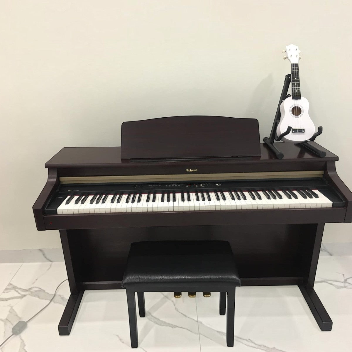Đàn piano điện Roland HP3 dáng tủ màu gỗ tối tại Tân Nhạc Cụ, kèm ghế đàn và đàn ukulele trắng đặt trên giá