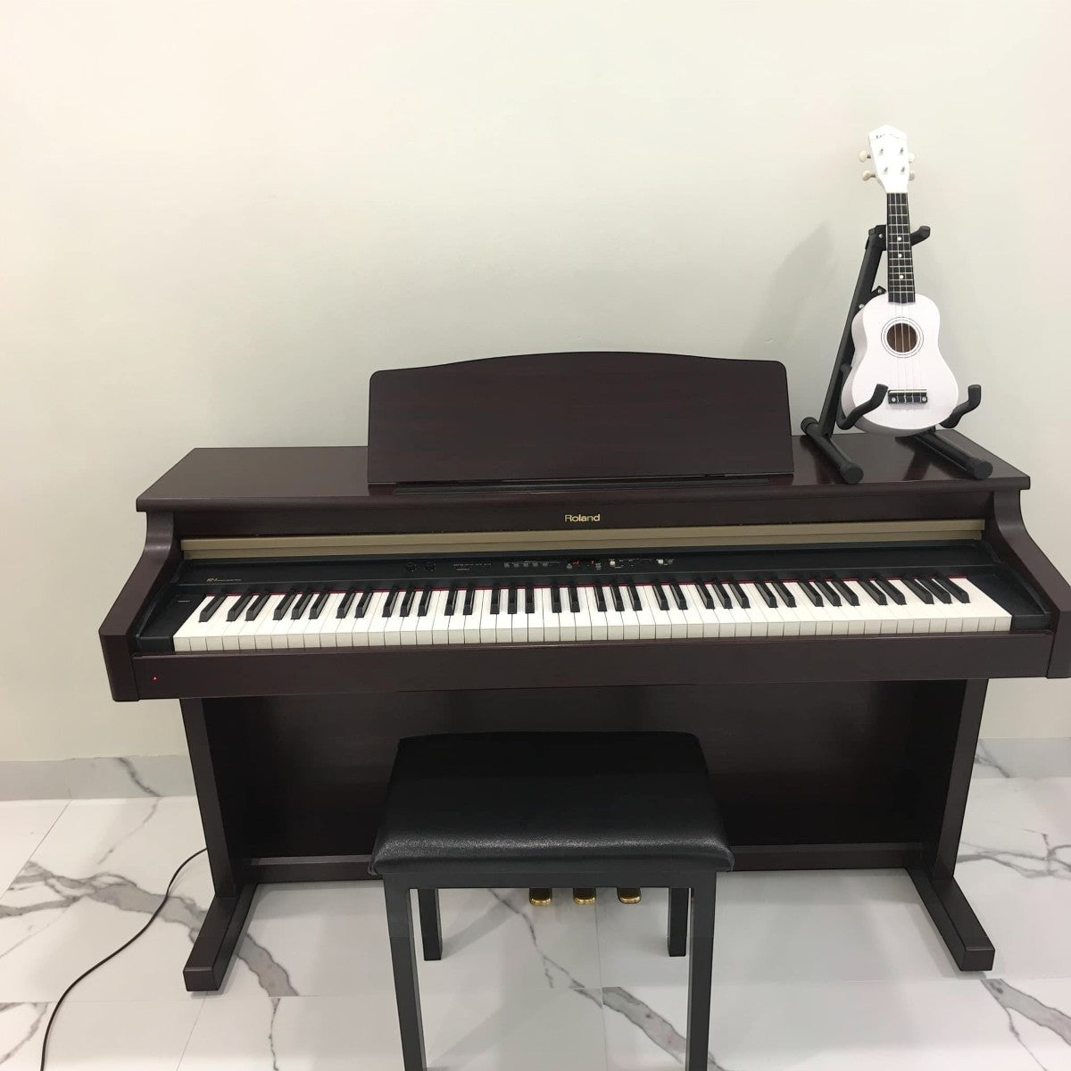 Đàn piano điện Roland HP3 dáng tủ màu nâu gỗ sang trọng tại Tân Nhạc Cụ, kèm ghế đàn và đàn ukulele trắng bên cạnh.